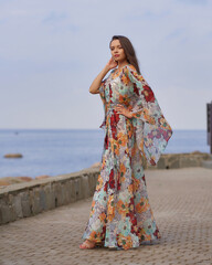 Pretty girl wearing colorful summer dress and walking at embankment with view on the sea