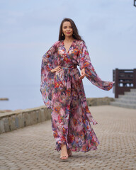 Young fashionable woman in colorful pnk dress with floral design walking at city embanlment with...