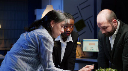 Diverse businesspeople coworkers standing at conference table analyzing financial graphs paperwork late at night. Multi ethnic teamwork planning company presentation in meeting room