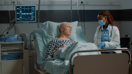African american medic talking to sick patient in hospital ward with nasal oxygen tube and pulse rate monitor. Old man getting medical advice from professional doctor with face mask