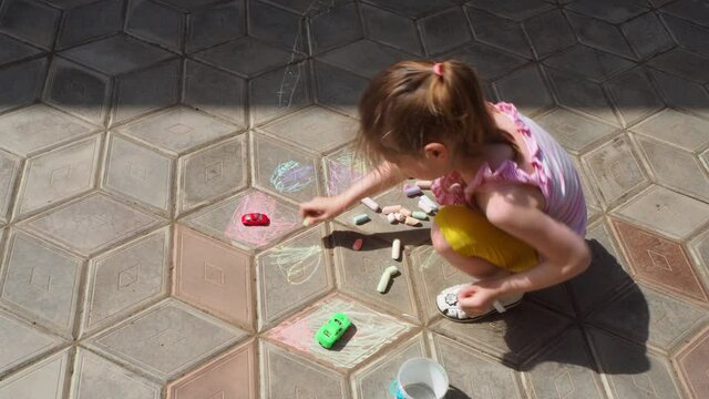 Girl draws with crayons on the paving stones in summer sunny day, top view