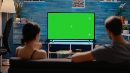Caucasian people watching green screen layout on tv display in living room at home. Young man and woman looking at copy space and chroma key for isolated template and mockup background