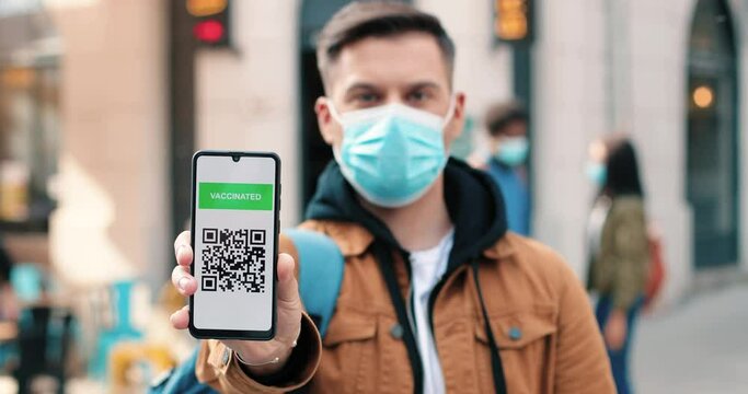 New normal. Portrait view of the serious man wearing protective mask looking at the camera and showing vaccine passport at his smartphone while standing at the empty street during the coronavirus