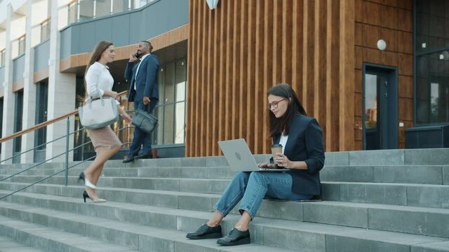 Attractive young woman is using laptop and drinking to go coffee sitting on stairs near modern business center working outdoors. People and urban lifestyle concept.