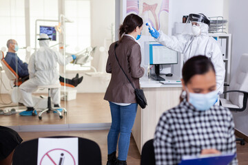 Stomatology receptionist using digital thermometer to measure patient temperature dressed in ppe suit as safety precaution during global outreabk with coronavirus. Dentist treating teeth.