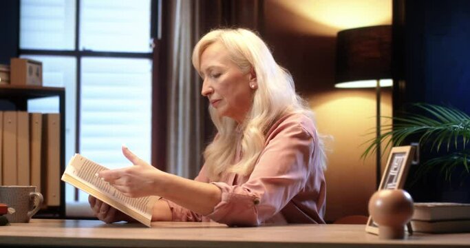 Beautiful gray-haired woman sitting at home and reading interesting book on background of warm floor lamp light.