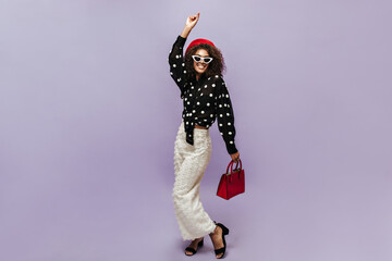 Positive cool girl with curly hair, sunglasses and modern beret in polka dot long sleeve blouse and white trousers smiling.