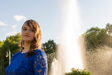 Pretty teen girl with makeup smiling and posing in the park. Female model with elegant hairstyle. Fountain in background