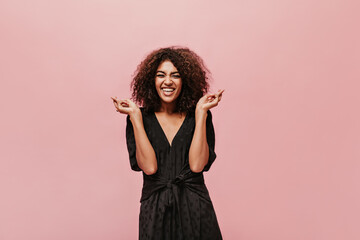 Cheerful girl with curly short hairstyle in black polka dot dress looking into camera, laughing and crossing her fingers.