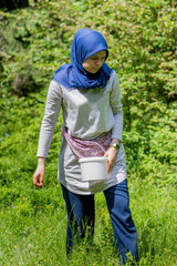 Muslim girl walking on the meadow with plastic bucket for collecting blueberries. Female worker picking organic wild bilberries on the mountain