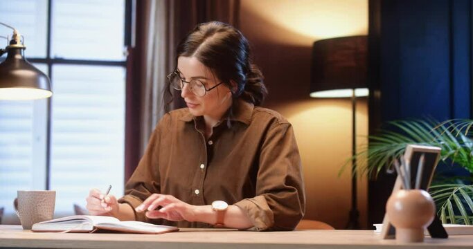 Beautiful stylish woman with glasses sitting at home at desk thinking and writing down her ideas in notebook.