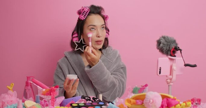 Asian woman applies face powder with cosmetic brush talks to followers wears hair rollers grey bathrobe sits at table in front of smartphone camera records livestreaming video isolated on pink