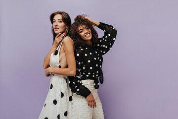 Stylish cool girl with silver earrings in polka dot light dress looking into camera and posign together with wavy haired woman in long sleeve blouse.