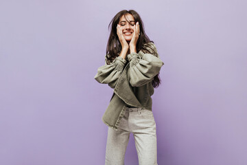 Charming stylish girl with long dark hair in long sleeve olive jacket and modern light pants smiling with closed eyes on lilac backdrop.