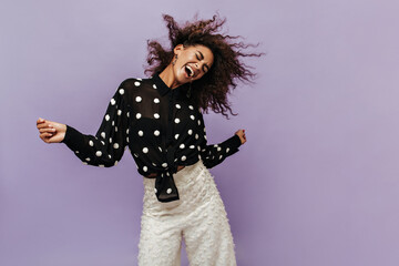 Emotional cool girl in fashionable black shirt and white trousers playing her curly hair and laughing with closed eyes on lilac backdrop.