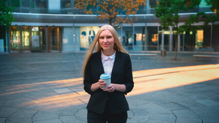 Elegant blond woman in suit and with long hair drinking coffee from white cup in patio with trees.