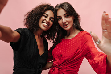 Stylish curly haired girl in dark clothes smiling, winking and taking selfie with cool friend with earrings in red dress.