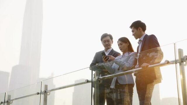 team of asian business people talking looking at cellphone outdoors on rooftop in downtown financial district of modern city