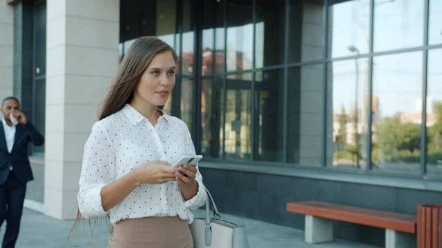 Dolly shot of ambitious young businesswoman walking along modern office center using smartphone texting concentrated on internet communication