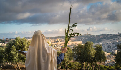 Fototapeta premium Succot (Feast of Tabernacles) in Jerusalem: Jewish man in a Tallit praying while waving the Four Species, with a view towards the Temple Mount, the Old City and the Mount of Olives