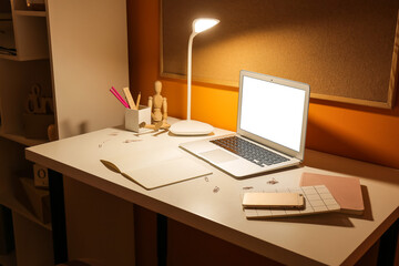 Stylish workplace with glowing lamp and modern laptop near color wall at night