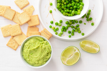 Bowl with tasty green pea hummus, lime and crackers on light wooden background