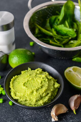 Bowl with tasty green pea hummus, lime, garlic and spinach on dark background, closeup