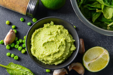 Bowl with tasty green pea hummus, lime and garlic on dark background, closeup