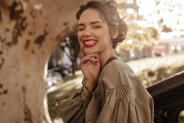 Positive girl with wavy hair and bright lipstick smiling sincerely outside. Trendy lady in denim clothes winking outdoors..
