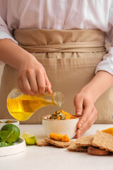 Woman pouring oil from jug into bowl with tasty carrot hummus on light table, closeup
