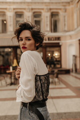 Fototapeta premium Short-haired woman in white blouse with long sleeve and dark lace poses in city. Girl with curly hair and red lips looking into camera at street..