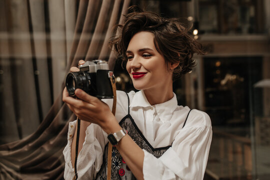 Wavy-haired girl with red lipa in light blouse holding camera in cafe. Stylish woman with brunette hair making photo inside..