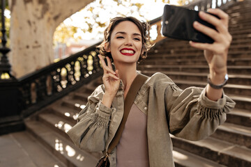 Joyful woman in olive denim jacket showing peace sign and smiling outside. Short-haired girl with red lips making selfie on backdrop of stairs..
