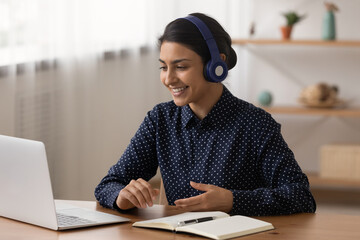 Smiling Indian woman in wireless headphones using laptop, making video call, student involved in...