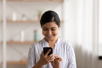 Head shot smiling Indian woman using smartphone, having fun with device, standing at home, positive...