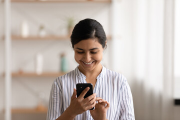 Fototapeta premium Head shot smiling Indian woman using smartphone, having fun with device, standing at home, positive attractive young female typing writing message in social network, chatting or shopping online