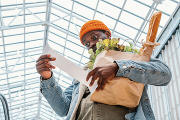 Surprised African-American man in denim jacket looks at receipt total in sales check holding paper...