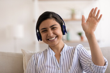 Head shot portrait of smiling Indian millennial woman in headphones waving hand, sitting on couch, young female involved in internet meeting, video call, happy mentor teacher leading online lesson