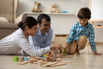 Fototapeta na wymiar Happy Indian young family with little son playing with toys, wooden blocks and dinosaurs on warm wooden floor with underfloor heating at home, smiling mother, father and preschool kid having fun