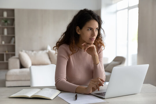 Pensive young Hispanic woman sit at desk at home office work online on laptop thinking pondering. Thoughtful millennial Latino female use computer look in distance make plans or solve problem.