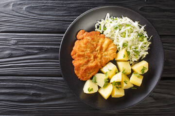 Kotlet Schabowy Polish Breaded Pork with boiled potatoes and cabbage salad close-up in a plate on a black wooden table. Horizontal top view from above - obrazy, fototapety, plakaty