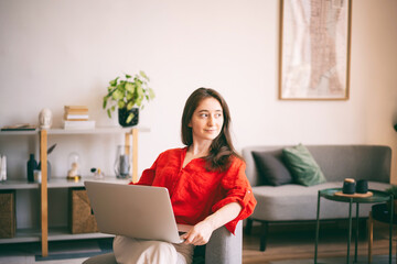 Portrait Young happy brunette woman in red shirt working on laptop sitting on chair at home in...