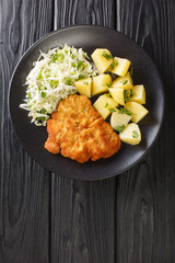 Kotlet Schabowy Polish Breaded Pork Chop with boiled potatoes and cabbage salad close-up in a plate on a black table. Vertical top view from above