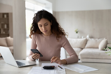 Young Hispanic woman sit at desk at home manage household finances paying bills calculating on machine cellphone. Millennial Latin female care of budget use smartphone for payment. Finance concept. - obrazy, fototapety, plakaty
