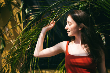 Portrait of a young happy brunette woman in a red t-shirt in profile with a leaf from a palm tree. Face without makeup, natural beauty trend