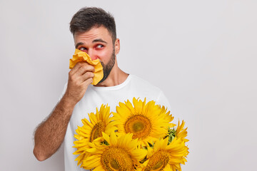 Handsome bearded man suffers from allergic rhinitis uses napkin to blow nose holds sunflower...