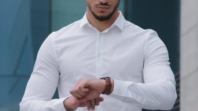 Cropped frame of unrecognizable hispanic arab man with beard wears formal white shirt stands in street outdoors looking at watch on hand hits dial with finger worries about being late time hour wait