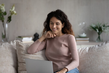 Happy millennial Latino woman relax on sofa at home watch webinar on laptop online. Smiling young...