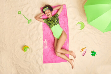 Obraz na płótnie Canvas Cheerful dark skinned woman with curly hair keeps handsbehind head wears green swisuit poses on pink towel near parasol sand toys drinks energetic beverage enjoys summer holidays. Rest concept