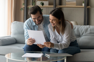 Smiling husband and wife sit on sofa reading received formal letter from bank about debt repayment, loan mortgage offer, reviewing together agreement, got invitation notice, health insurance concept